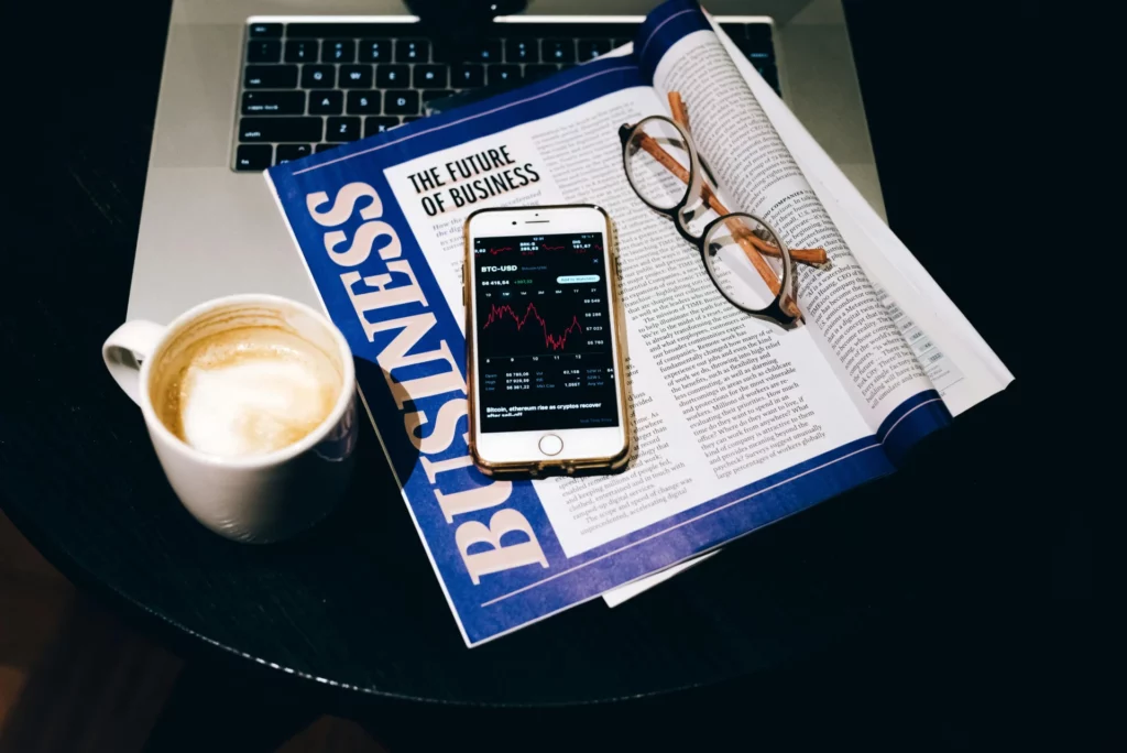 A smartphone displaying a crypto chart sits on a "Business" magazine with glasses, next to a coffee cup and laptop on a table representing news events in forex trading.