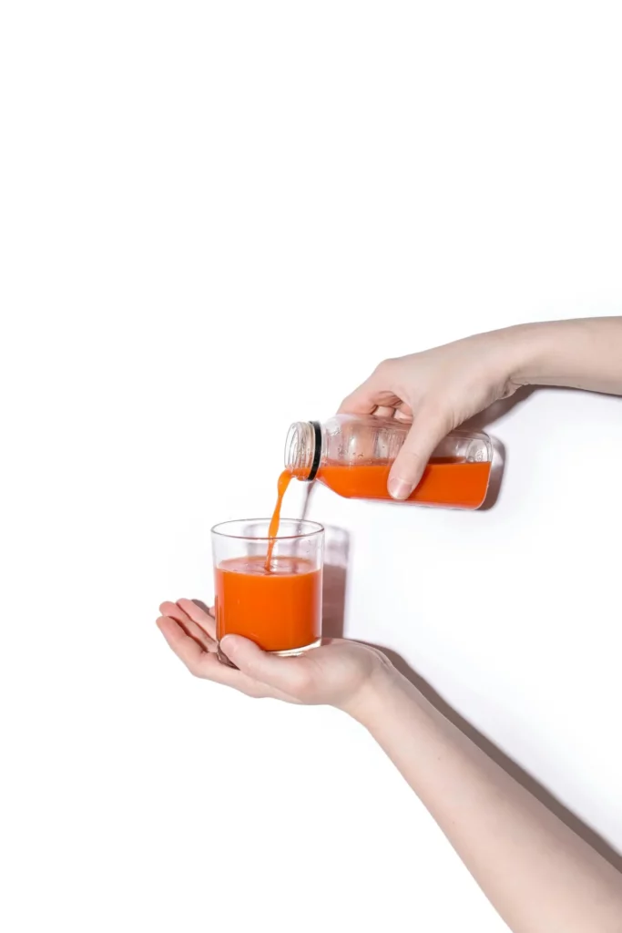 A hand pouring orange liquid from a bottle into a glass held by another hand, both set against a plain white background.