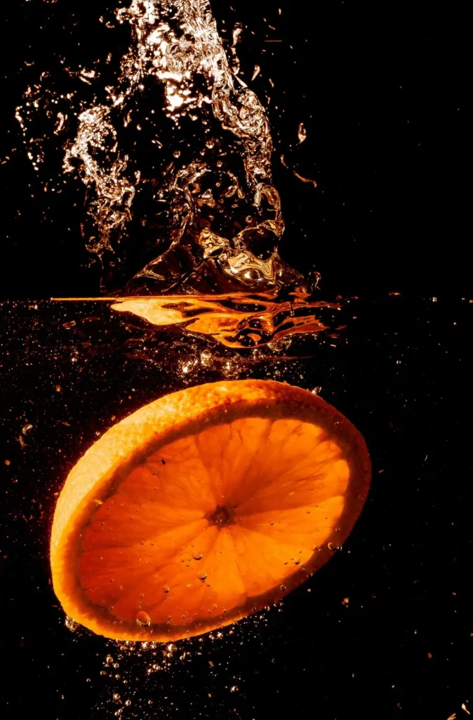 The image depicts a cross-section of an orange slice submerged in dark liquid, with water splashing and bubbling around it, creating a dynamic effect against a black background.