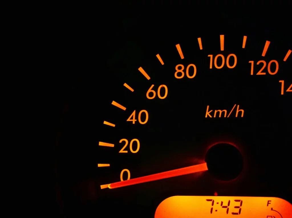 The image shows a car's speedometer illuminated in orange, with the needle pointing at 0 km/h.