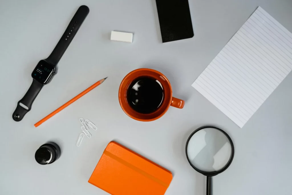 Minimal desk flat lay: orange coffee mug, orange notebook, black smartwatch, pencil, paperclips, eraser, and magnifying glass on gray background.