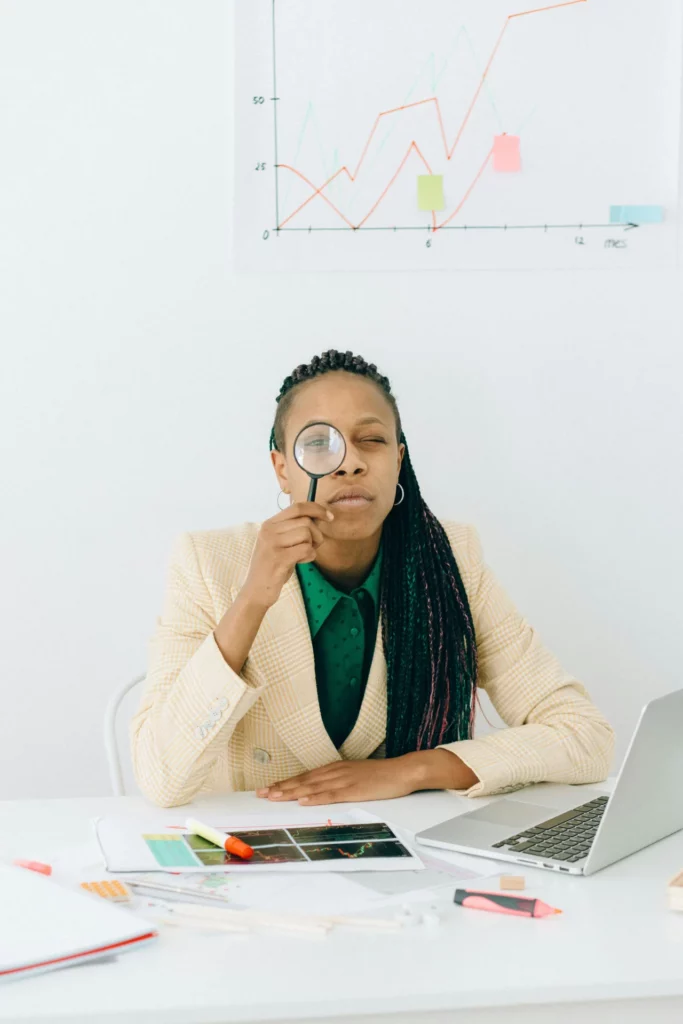 A skeptical woman with braids holds a magnifying glass to her eye, scrutinizing data at a desk covered in charts, highlighters, and a laptop, with a large annotated graph on the wall behind her.