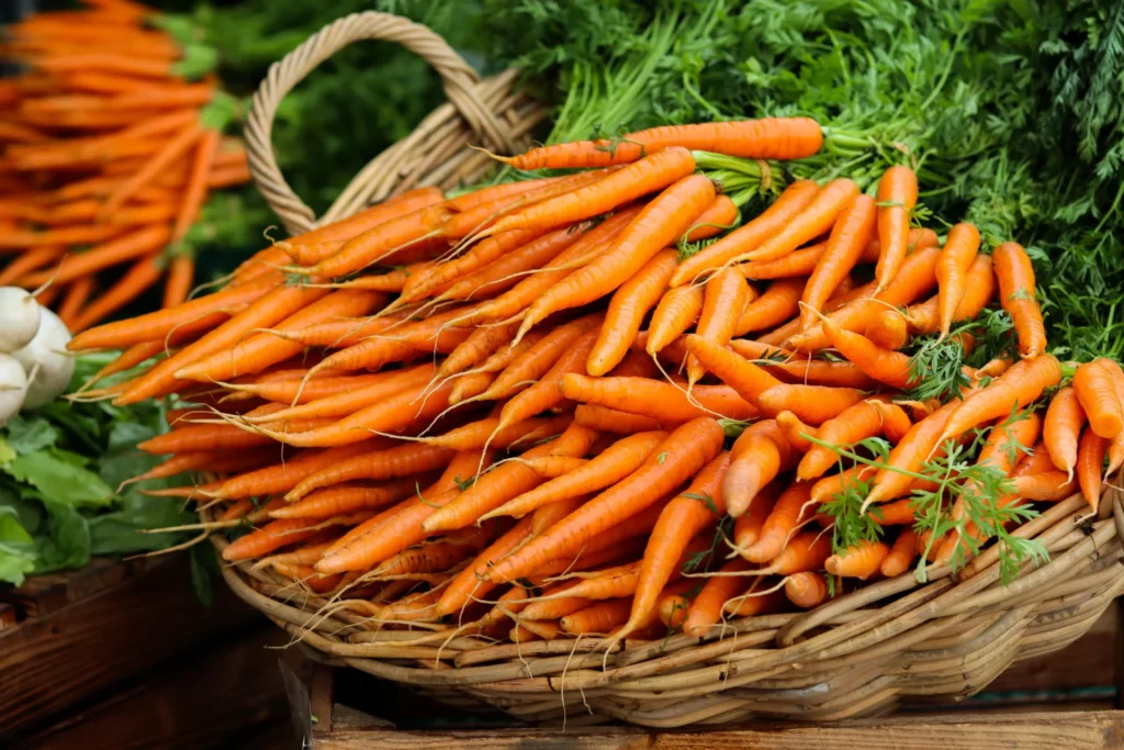 A wicker basket overflowing with fresh, bright orange carrots with green tops, displayed at a market.