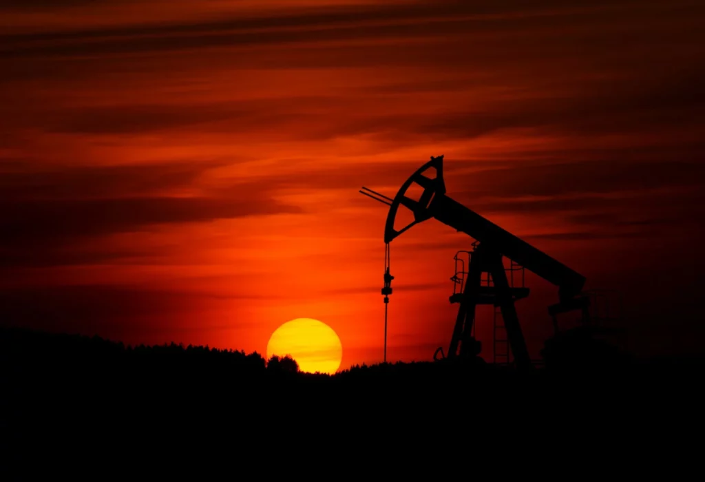 Silhouette of an oil pump jack against a vivid orange-red sunset sky, with the sun low on the horizon behind a dark tree line.