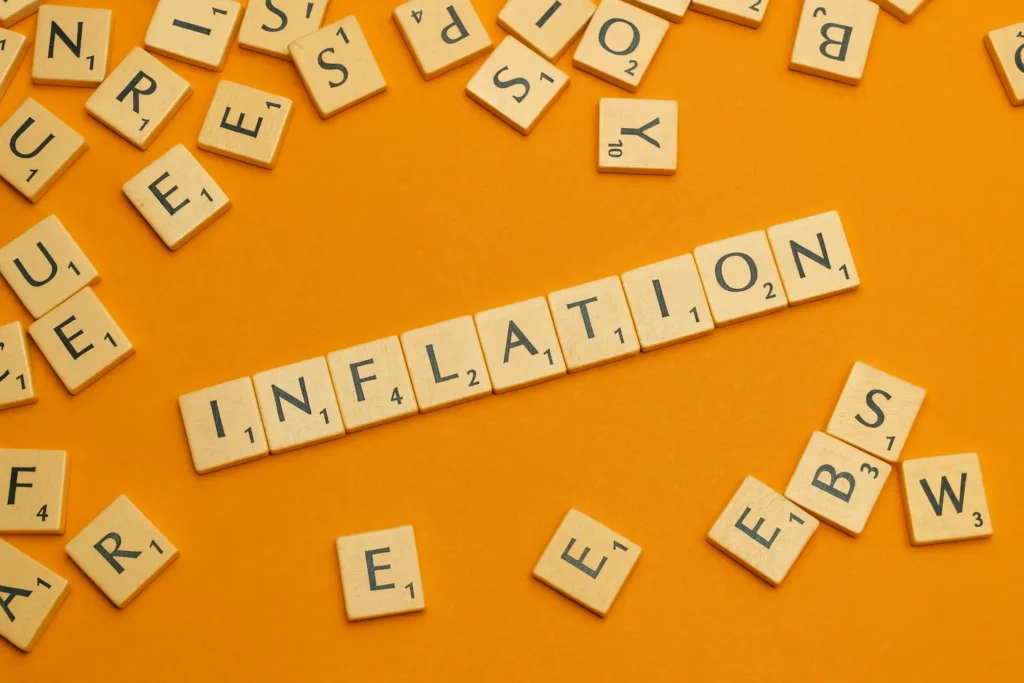 Scrabble tiles on an orange background spelling out "INFLATION" in a straight line, surrounded by scattered letter tiles.