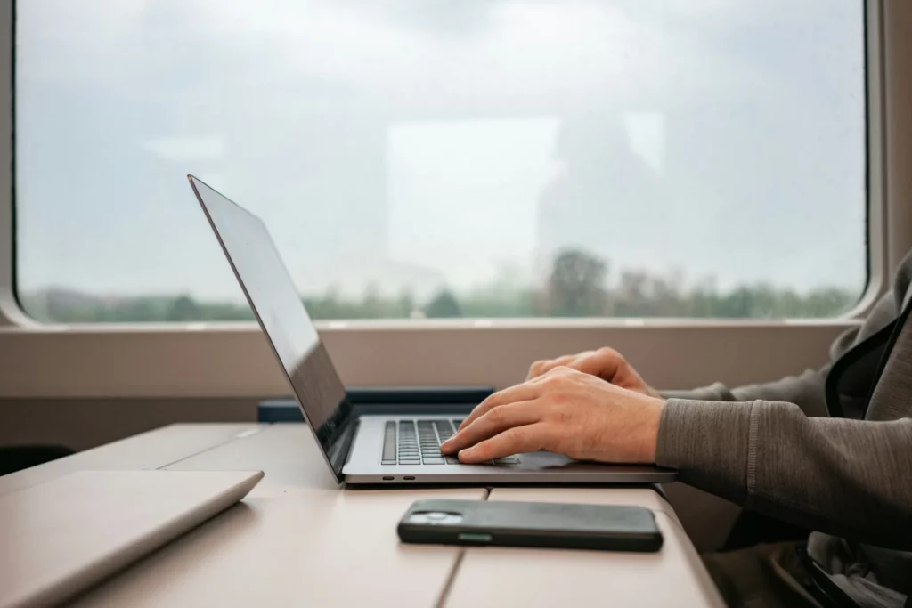 Person typing on a MacBook at a train table, phone and closed tablet beside it, blurred window view of passing scenery outside. Access instant funding prop firms from anywhere.