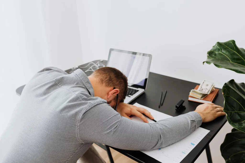 Exhausted trader slumped on desk showing loss of trading focus and consistency: head down on papers, laptop open, cash stack and plant nearby in a modern workspace.