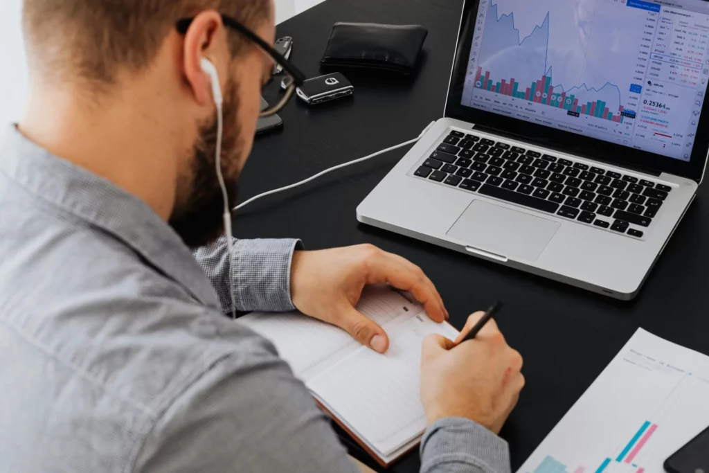 Man with glasses working at a desk with laptop showing financial charts, taking notes in a notebook, illustrating what are prediction markets and how people trade the future. 